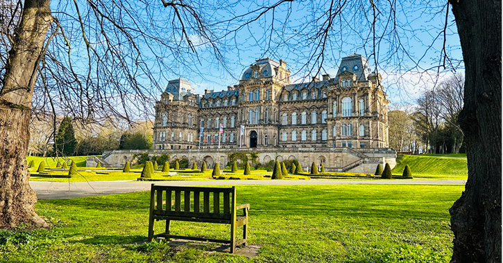 Exterior view of The Bowes Museum and grounds on a spring day with empty bench in foreground.